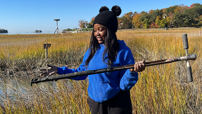 Precious Adesoji Adesoji ’23 M.S. standing in wetlands holding scientific equipment.