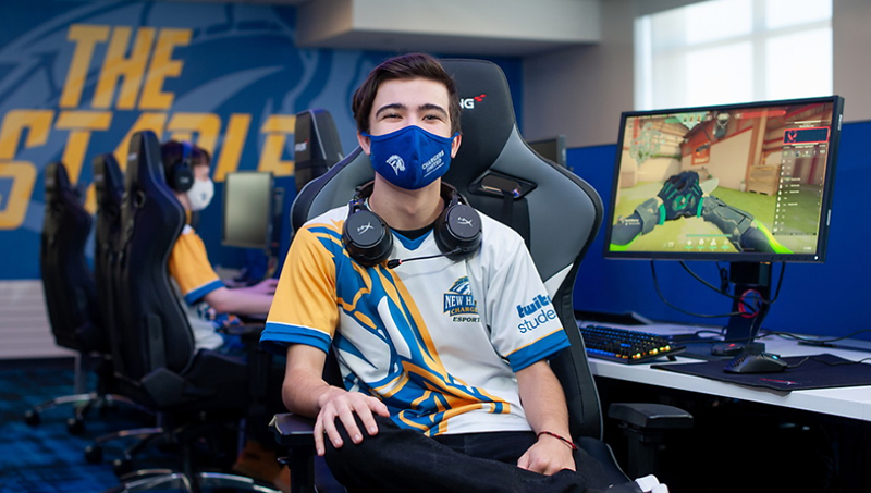 A student poses for a picture, in a gaming chair, in front of a student in the background playing a computer game.